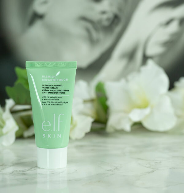A travel size tube of the e.l.f. Blemish Breakthrough Blemish Calming Water Cream, green with a white lid, standing in front of a dark background with white flowers