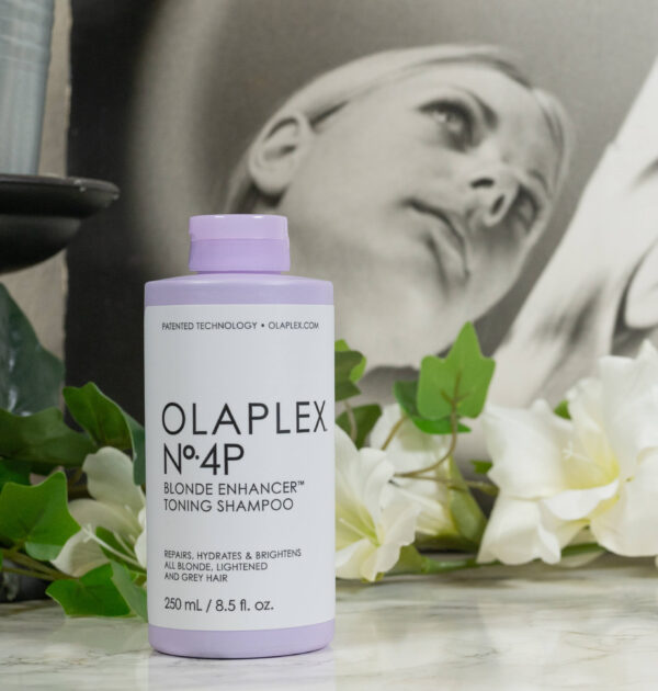 A bottle of Olaplex N° 4P Blond Enhancer Toning Shampoo in front of white flowers on a dark background