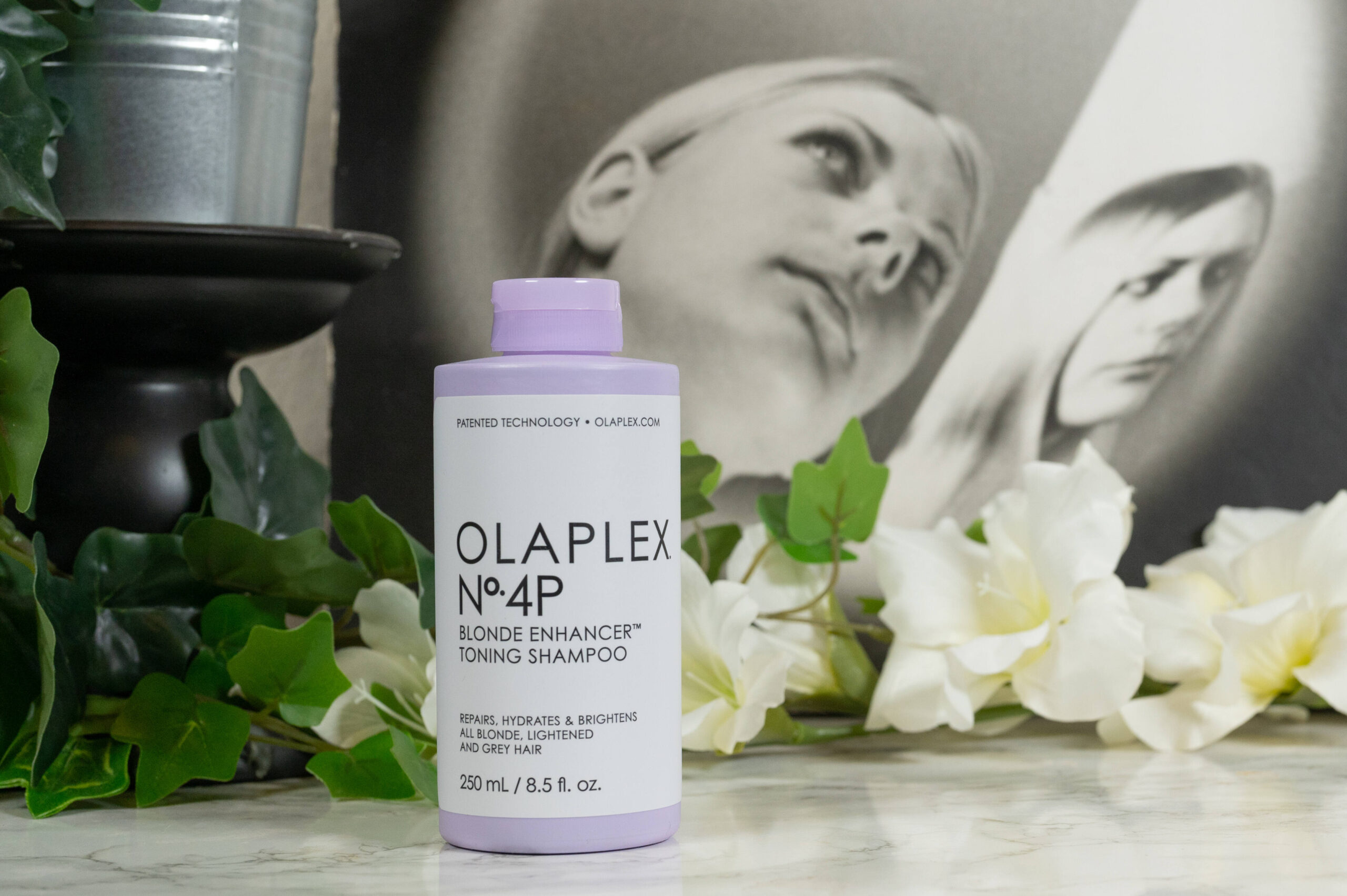 A bottle of Olaplex N° 4P Blond Enhancer Toning Shampoo in front of white flowers on a dark background