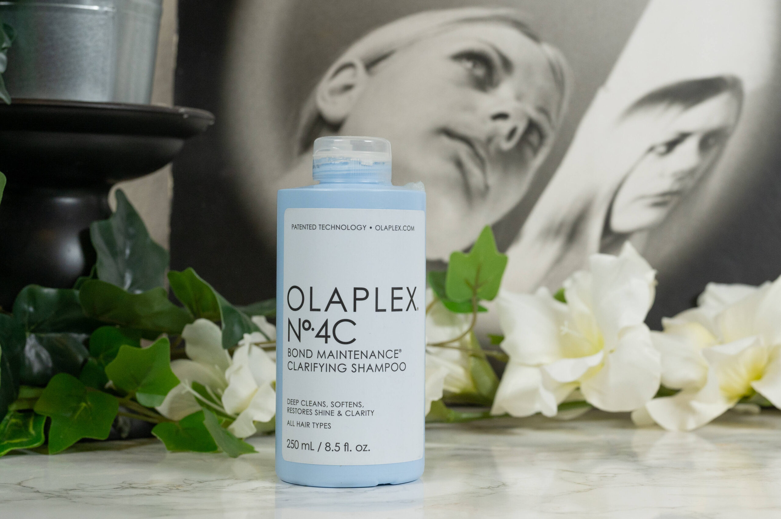 A bottle of Olaplex No 4C Bond Maintenance Clarifying Shampoo standing in front of white flowers on a dark background