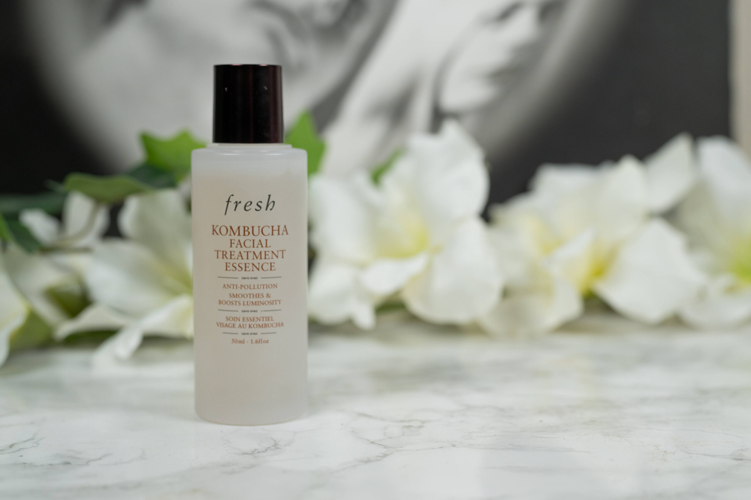 A small bottle of fresh Kombucha Facial Treatment Essence standing in front of white flowers on a dark background