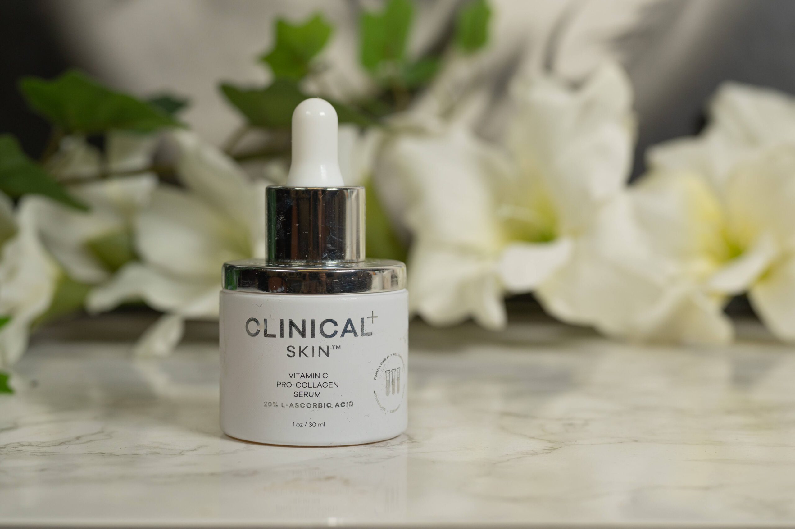 Clinical Skin Vitamin C Pro Collagen Serum standing in front of white flowers on a dark background. The bottle is white and opaque with a silver top.