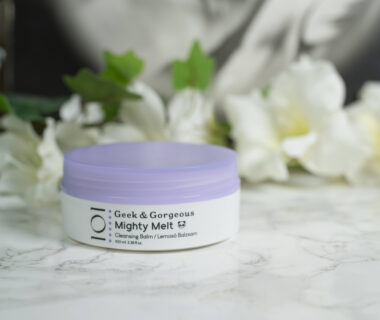 One pot of Geek and Gorgeous Mighty Melt Cleansing Balm, white with a lilac lid, standing in front of white flowers on a dark background