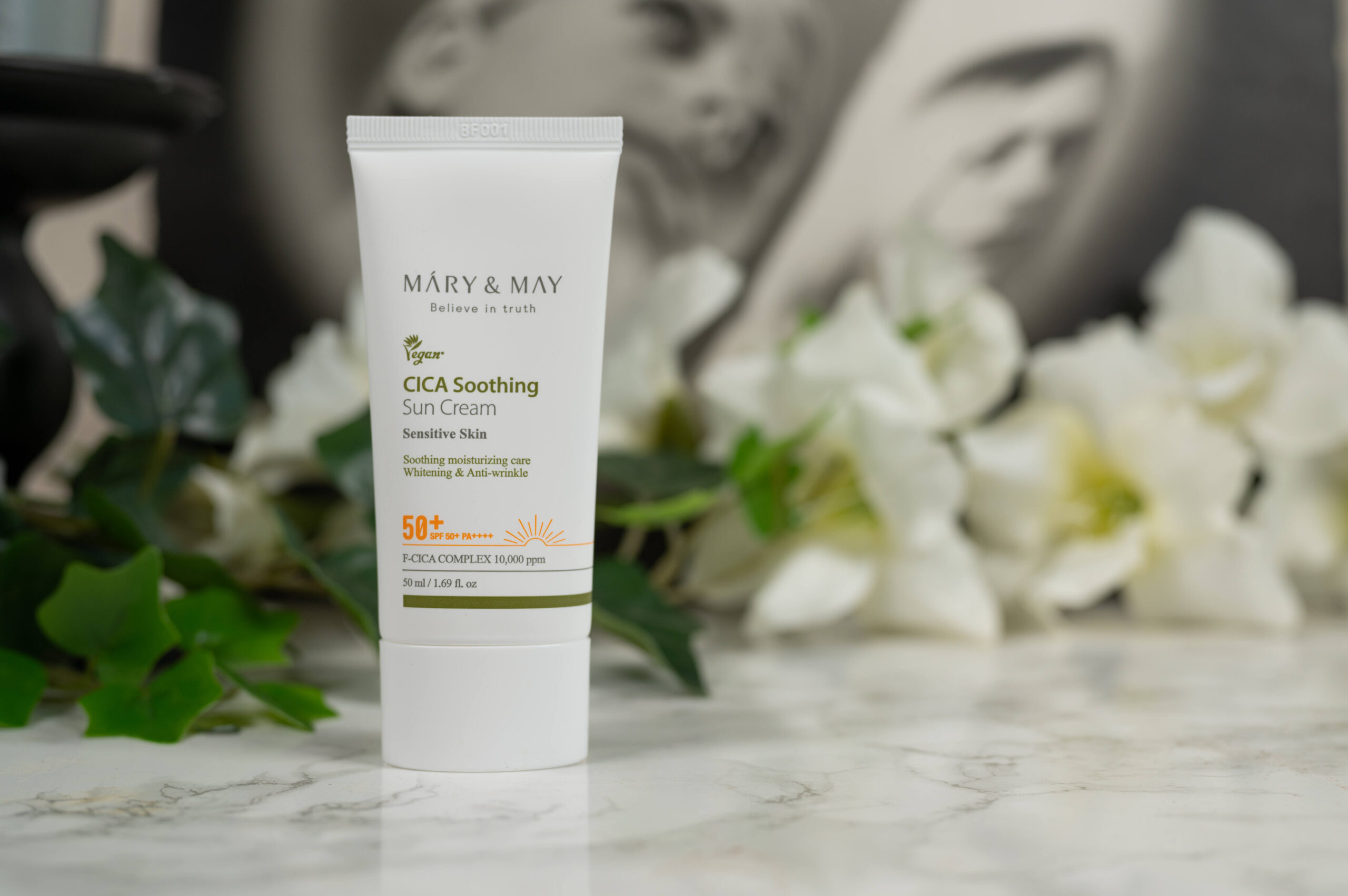A tube of Mary and May CICA Soothing Sun Cream SPF 50+ standing in front of a dark background with white flowers