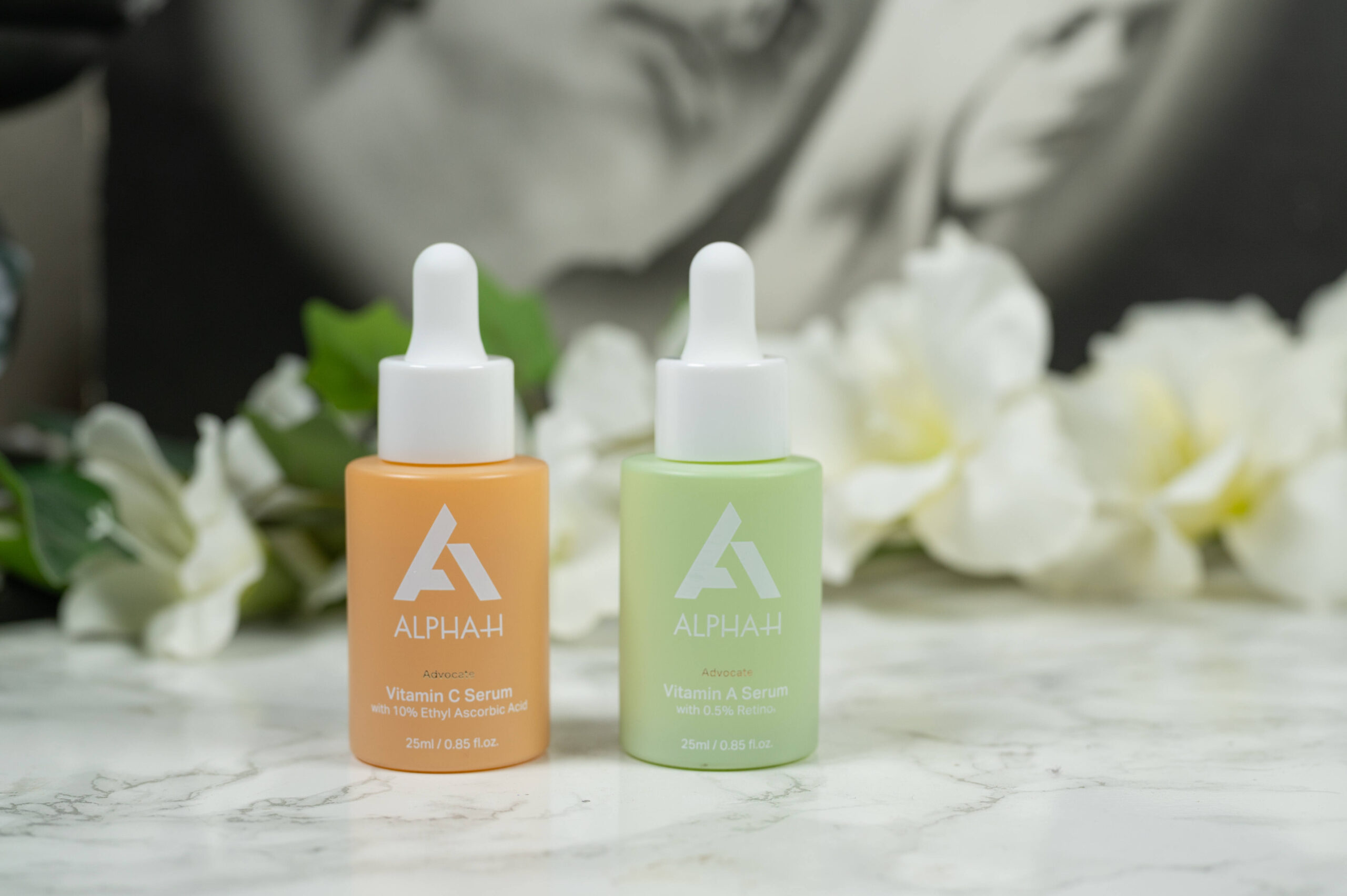 The Alpha-H Advocate Vitamin C and Vitamin A serum standing in front of a dark background with white flowers