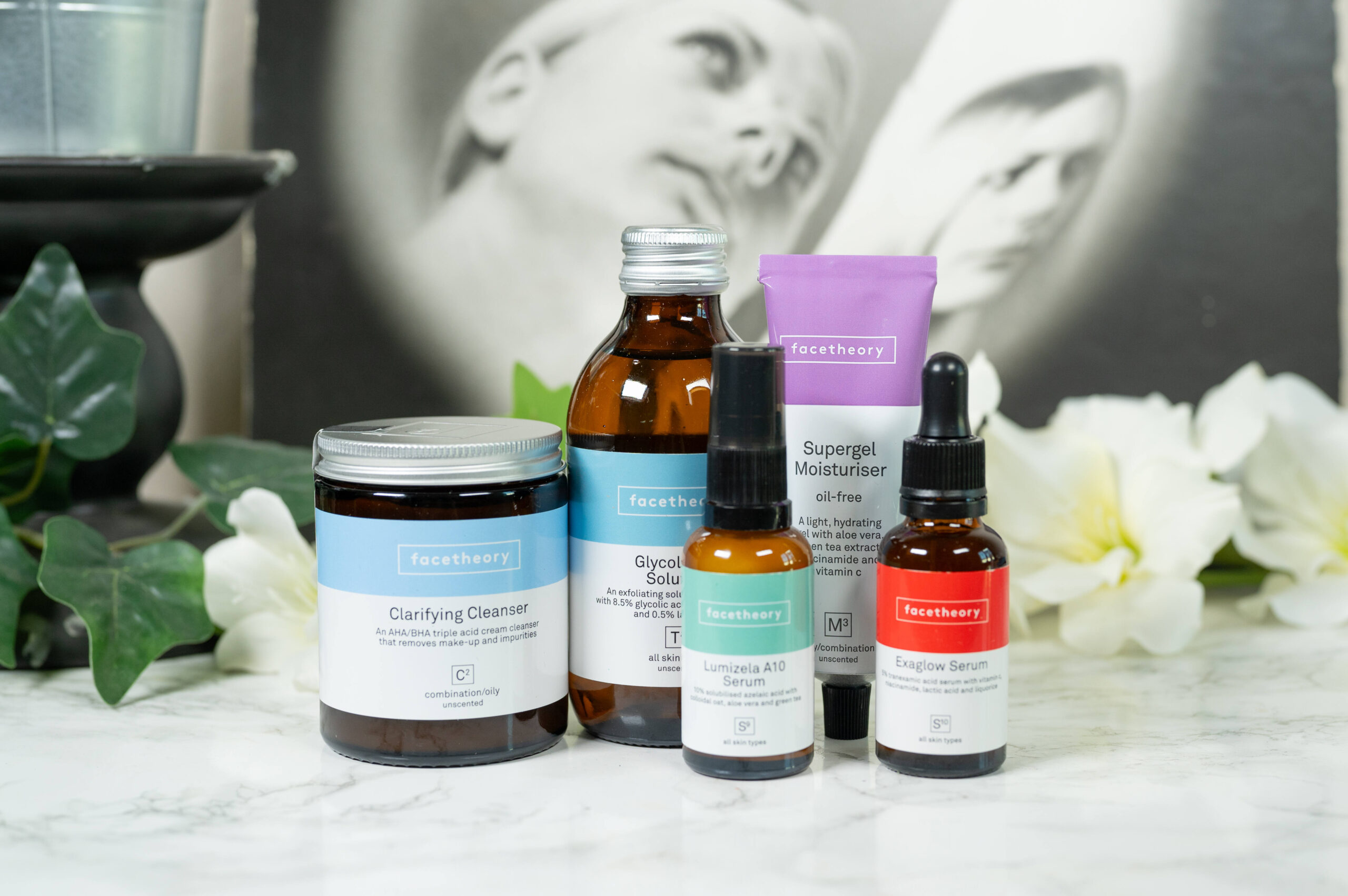 Facetheory Clarifying Cleanser, Glycolacsal Solution, Lumizela A 10 Serum, Exaglow Serum and Supergel Moisturizer standing in front of a dark background with white flowers
