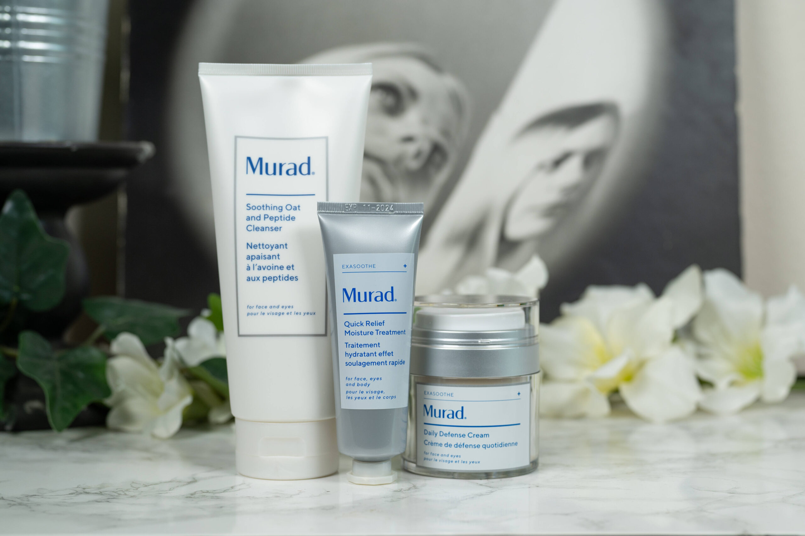 The Murad Soothing Oat and Peptide Cleanser, Quick Relief Moisture Treatment and Daily Defense Cream standing in front of a dark background with white flowers