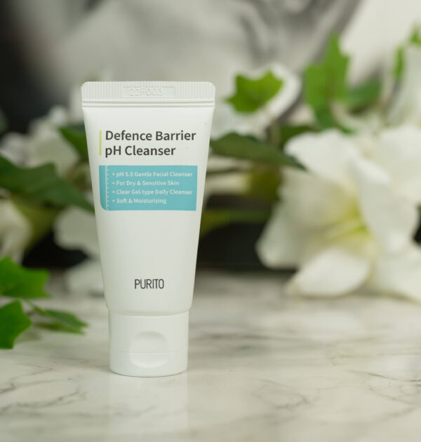 A travel size of the Purito Defence Barrier pH Cleanser standing in front of a dark background with white flowers