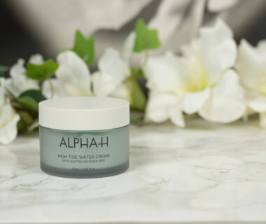 A pot of Alpha-H High Tide Water Cream standing in front of a dark background with white flowers.