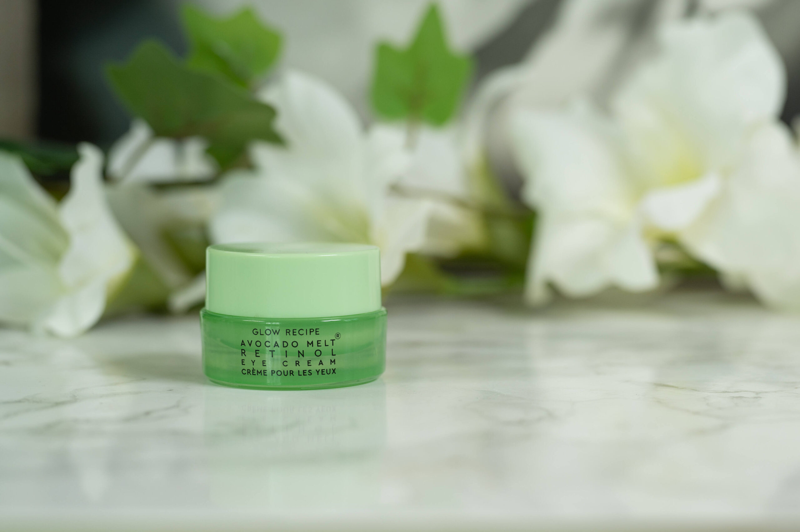 A travel size pot of Glow Recipe Avocado Melt Retinol Eye Cream standing in front of white flowers on a dark background