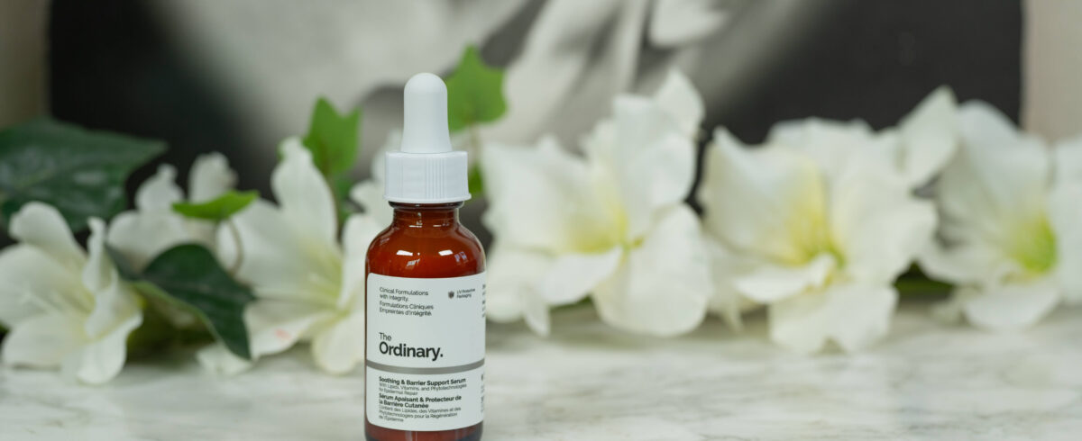 The Ordinary Soothing & Barrier Support Serum standing in front of a dark background with white flowers