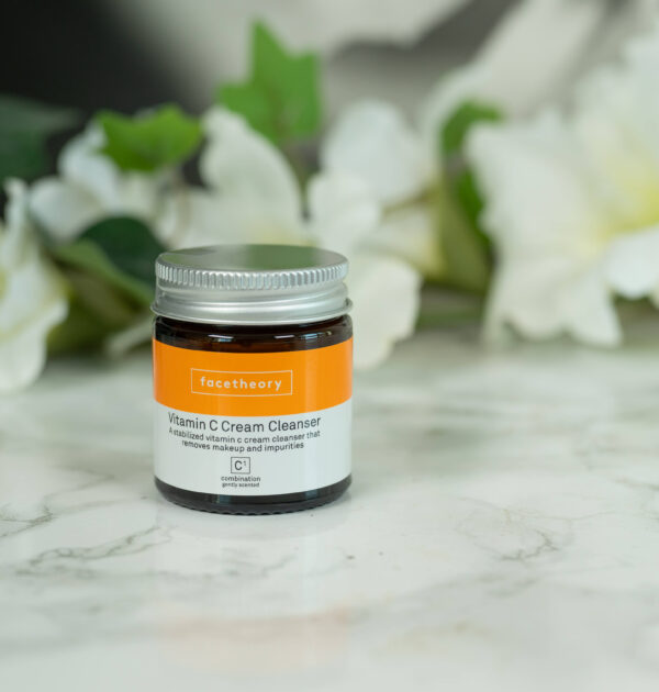 One pot of facetheory Vitamin C Cream Cleanser standing in front of a dark background with white flowers