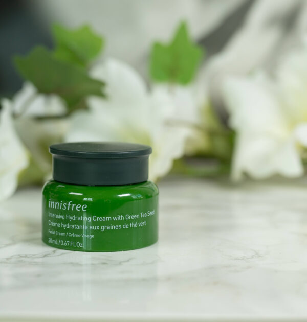A tube of innisfree Intensive Hydrating Cream with Green Tea Seed, a green pot, standing in front of a dark background with white flowers