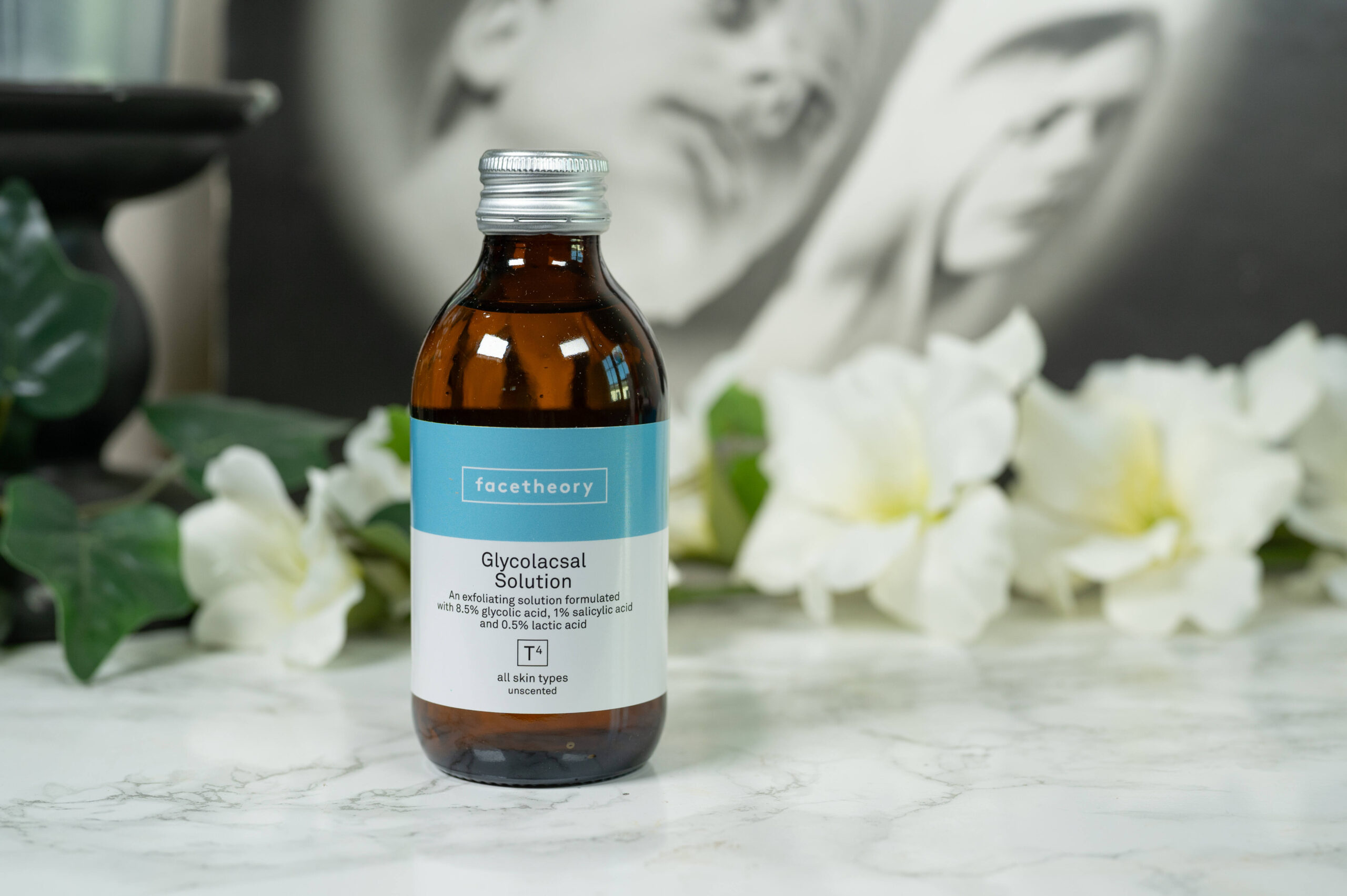 A bottle of facetheory Glycolacsal Solution (brown glass with a blue and white label) standing in front of a dark background with white flowers