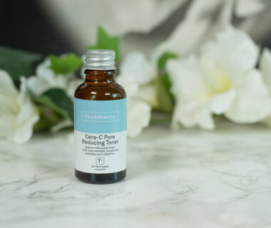 A travel size of the facetheory Cera-C Pore Reducing Toner standing in front of a dark background with white flowers