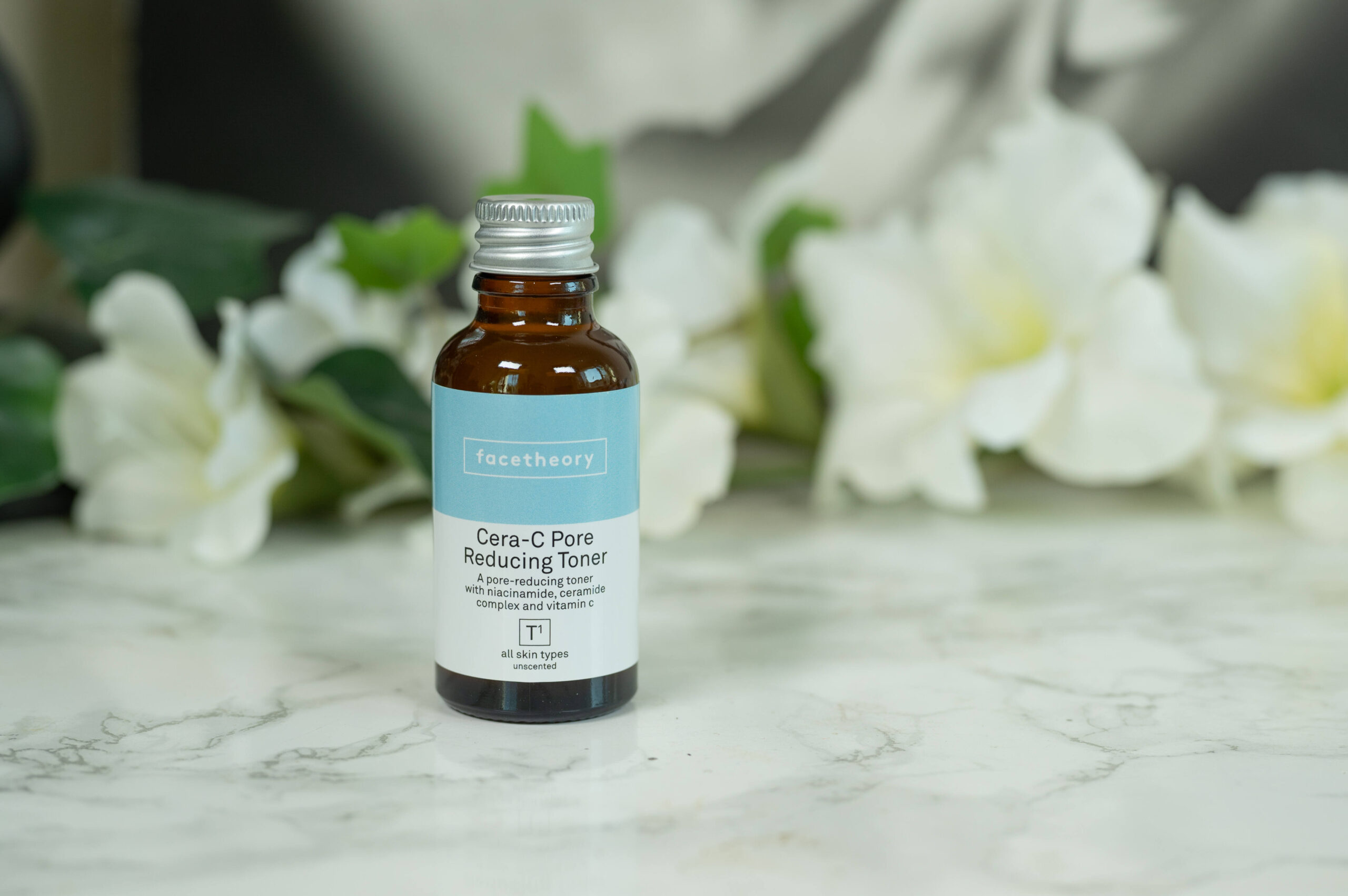A travel size of the facetheory Cera-C Pore Reducing Toner standing in front of a dark background with white flowers