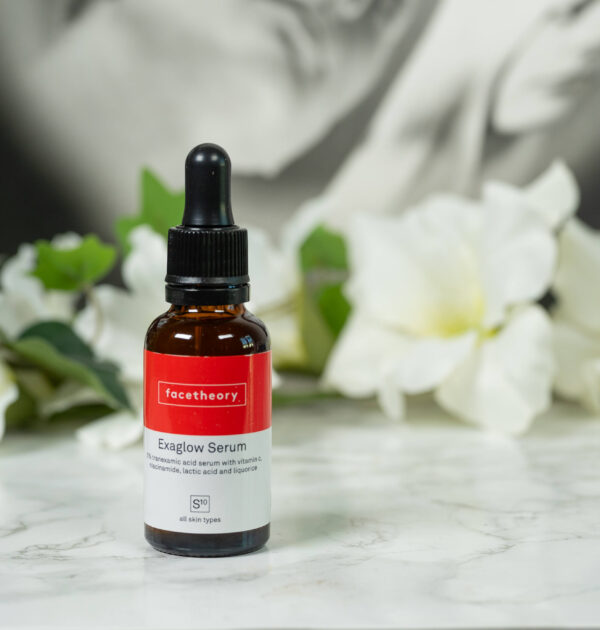 A bottle of Facetheory Exaglow Serum standing in front of a dark background with white flowers