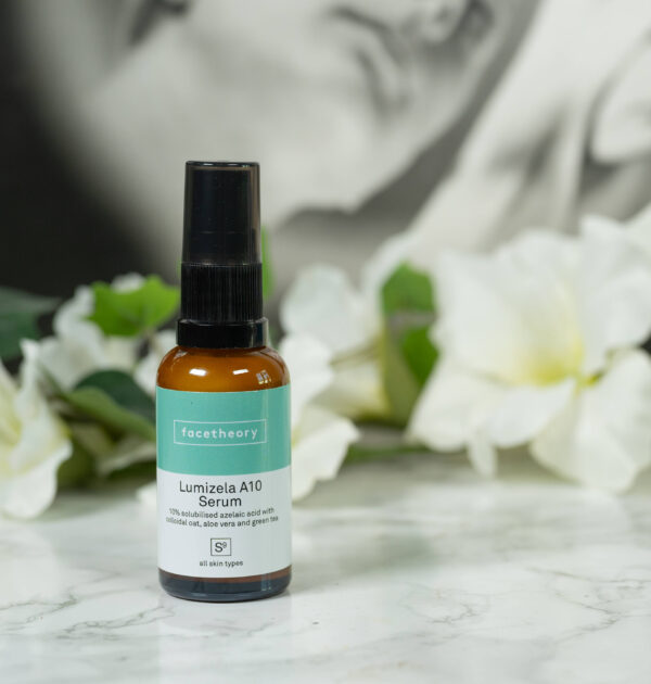 A bottle of Facetheory Lumizela A10 Serum standing in front of a dark background with white flowers