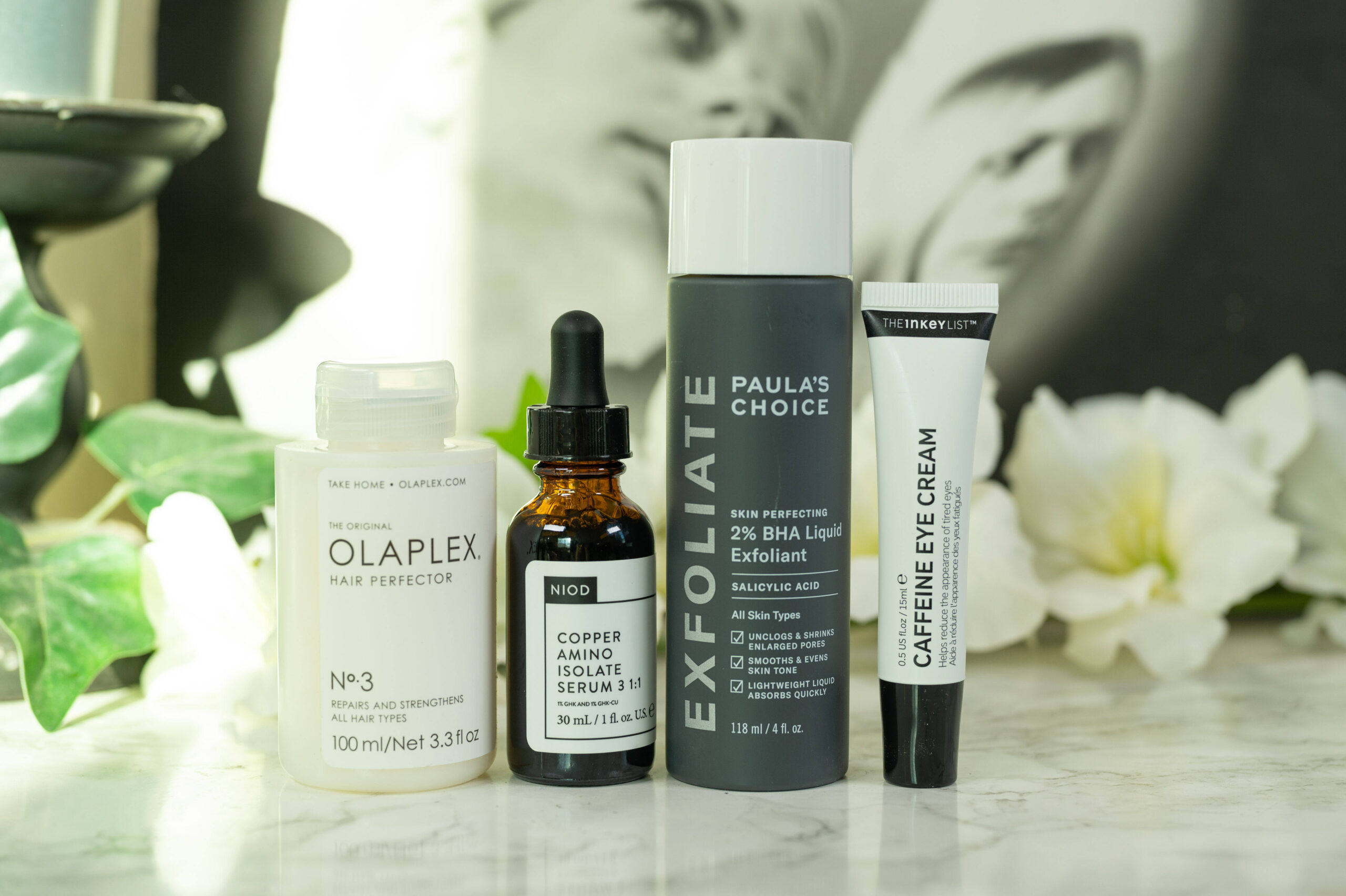 Skincare from Paula's Choice, The Inkey List and NIOD as well as hair care from Olaplex lined up in front of a dark background with white flowers