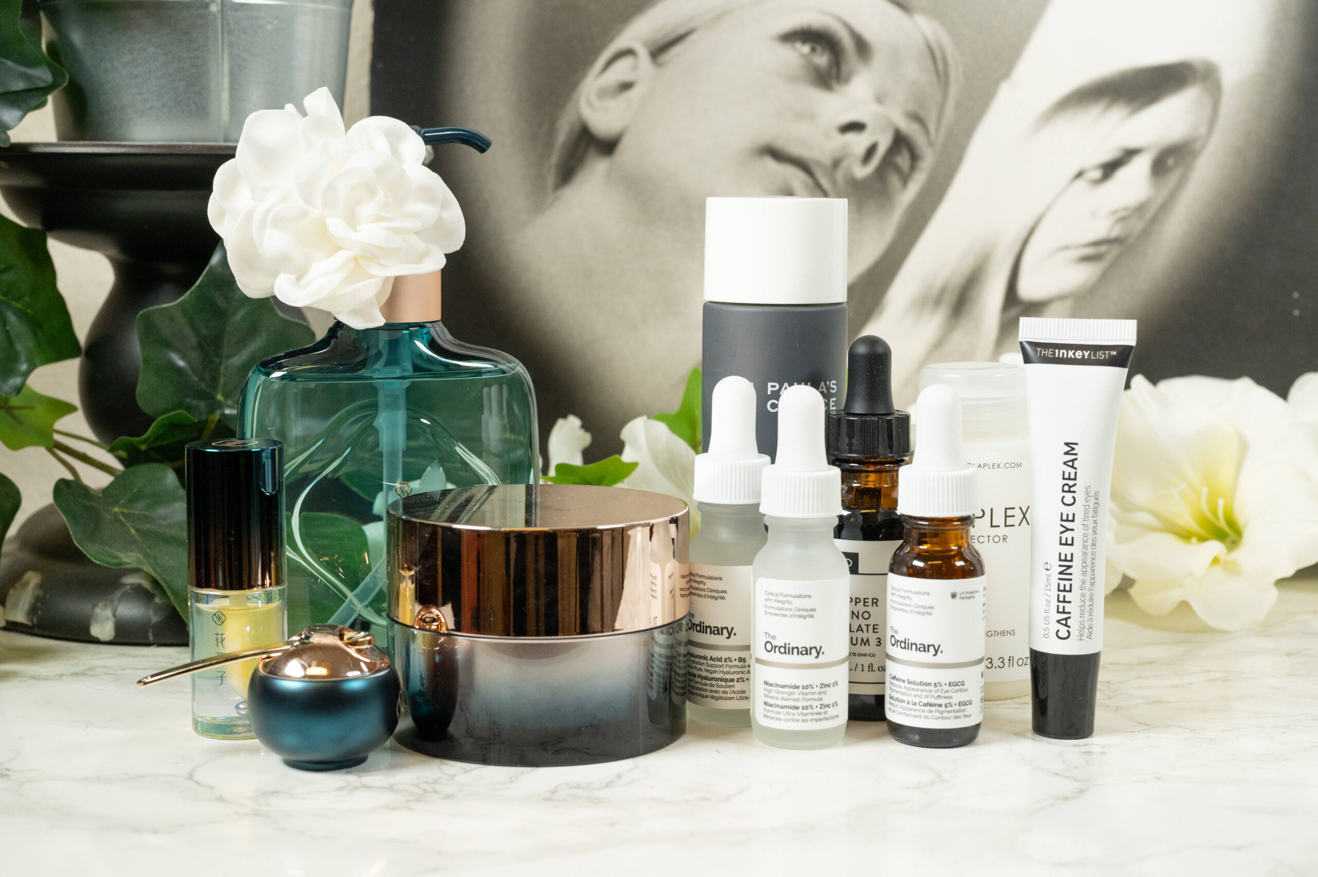 Skincare products from The Inkey List, Olaplex, Paula's Choice, The Ordinary and Florasis lined up in front of a dark background with white flowers
