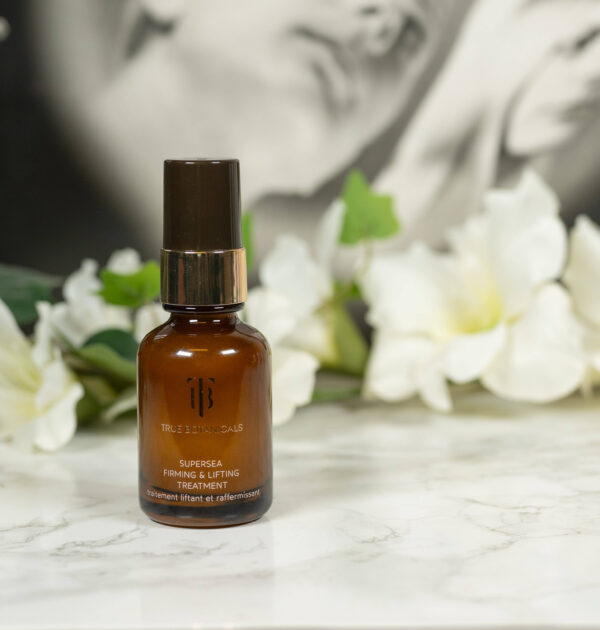 A bottle of the True Botanicals Supersea Firming & Lifting Treatment, made from brown glass, standing in front of a dark background with white flowers