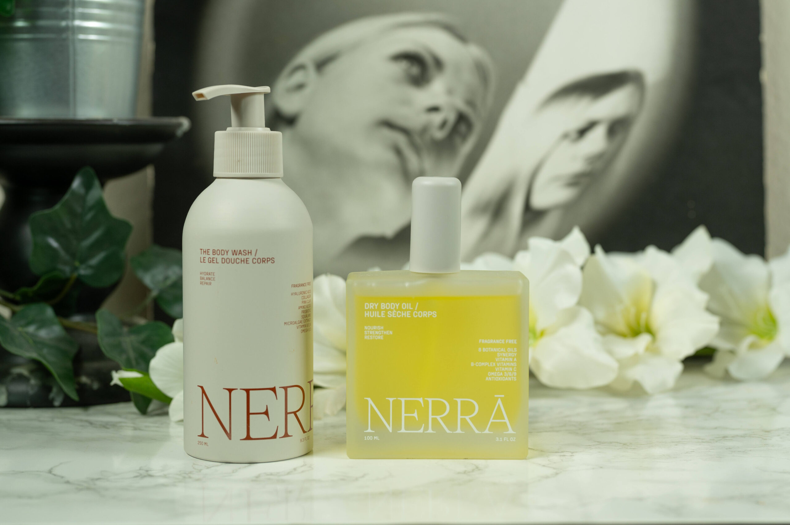 The Nerra Body Care Body Wash and Dry Body Oil standing in front of a dark background with white flowers