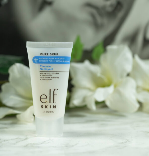 A travel size of the elf Pure Skin Cleanser standing in front of a dark background with white flowers.