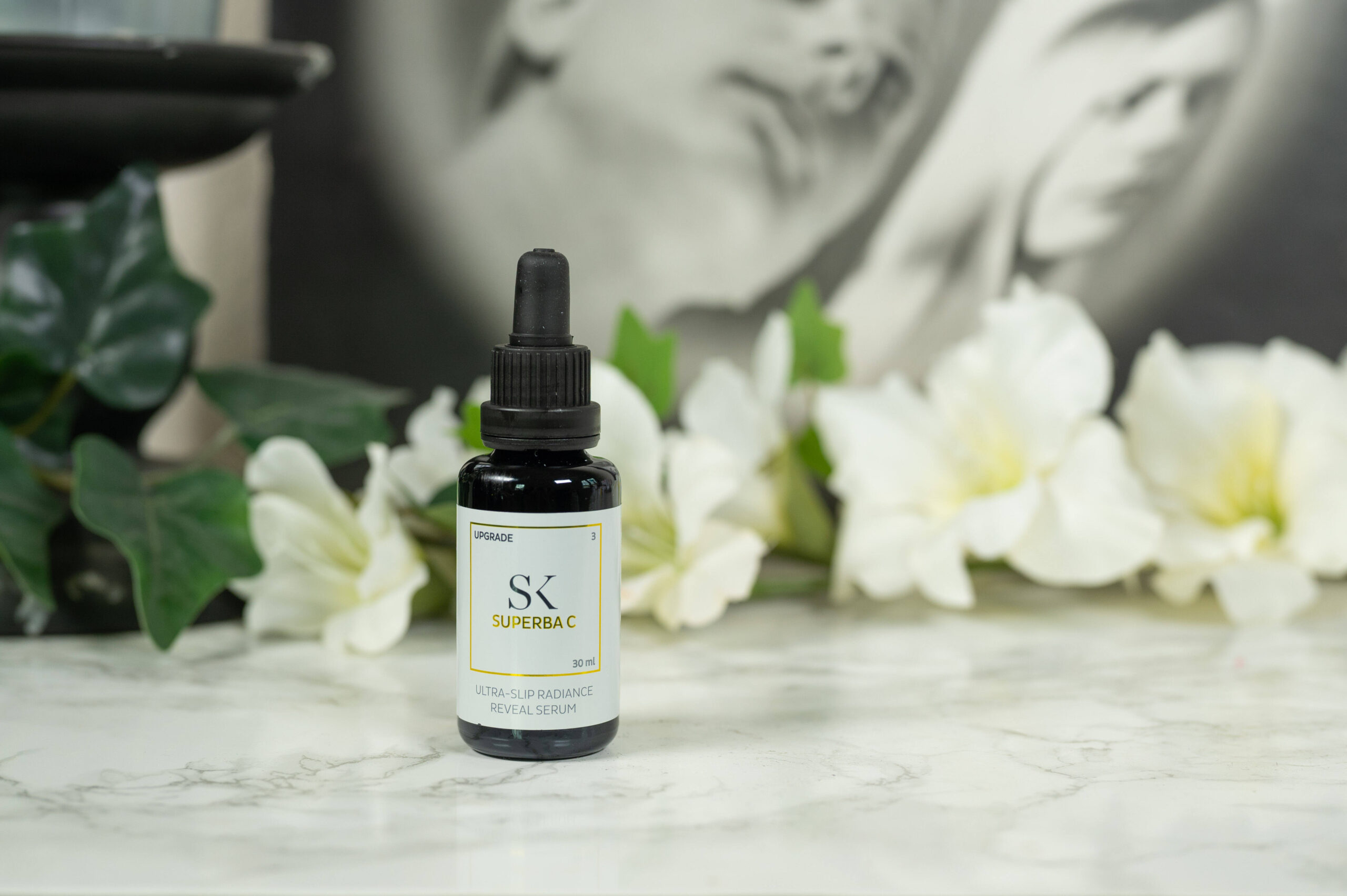A dark glass bottle with a dropper containing the Skintegra Superba C Serum standing in front of a dark background with white flowers