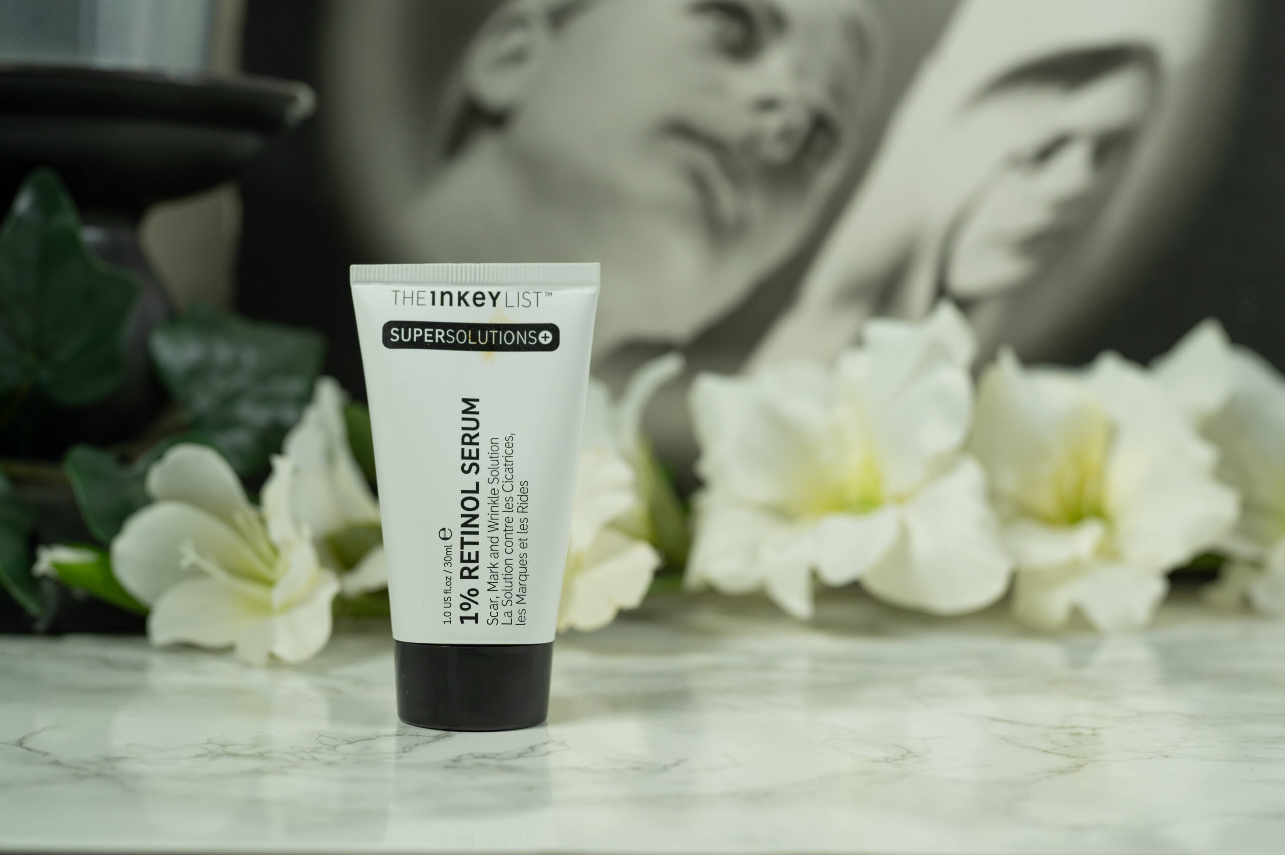 A tube of The Inkey List Scar, Mark and Wrinkle Solution standing in front of a dark background with white flowers
