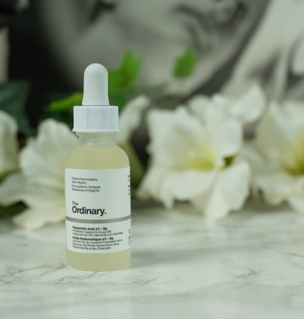 A bottle of the reformulated The Ordinary Hyaluronic Acid 2% + B5 Serum standing in front of a dark background with white flowers