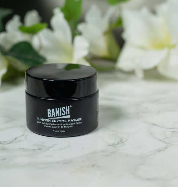 A pot of Banish Pumpkin Enzyme Mask standing in front of a dark background with white flowers