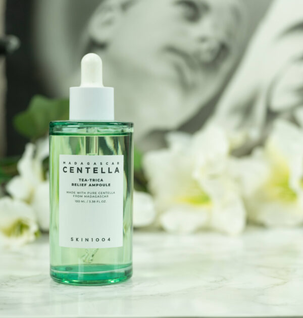 A bottle of SKIN1004 Tea-Trica Relief Ampoule standing in front of a dark background with white flowers