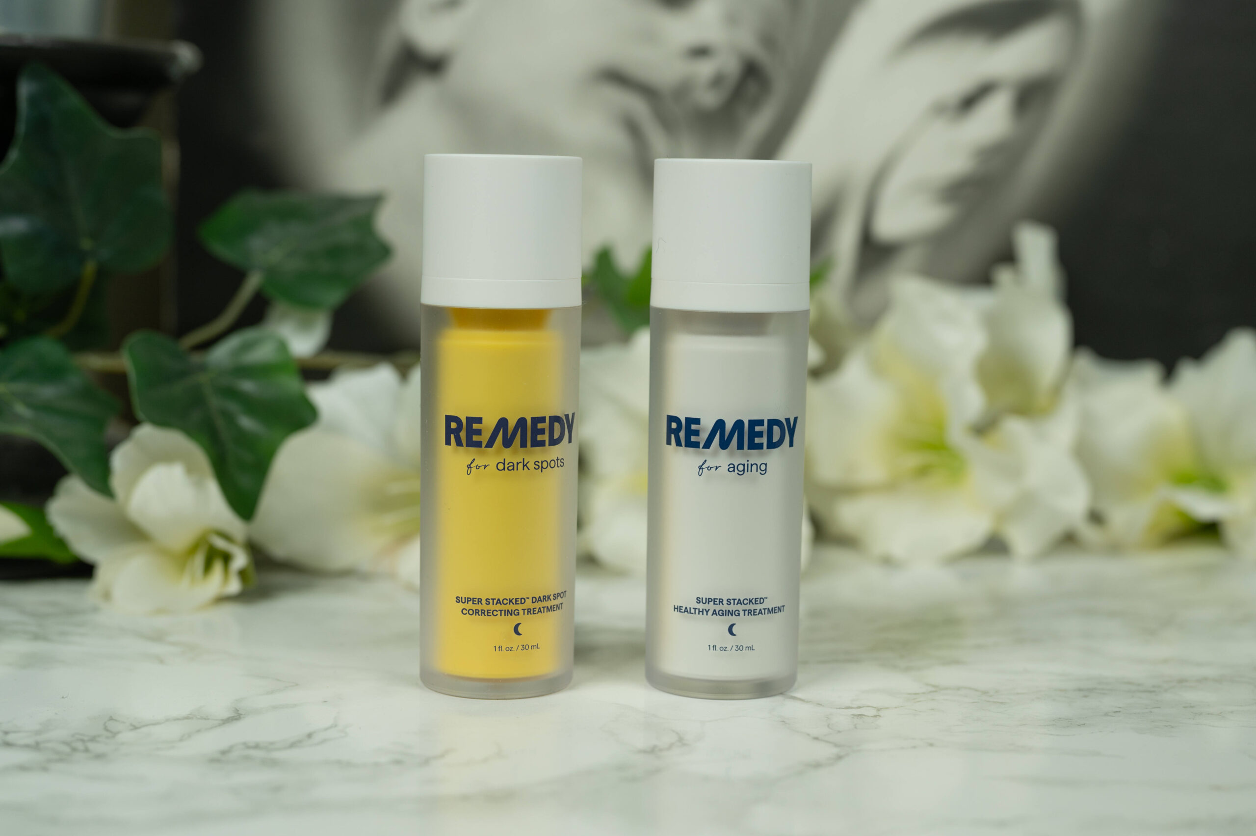Two bottles of Remedy skincare standing in front of a dark background with white flowers