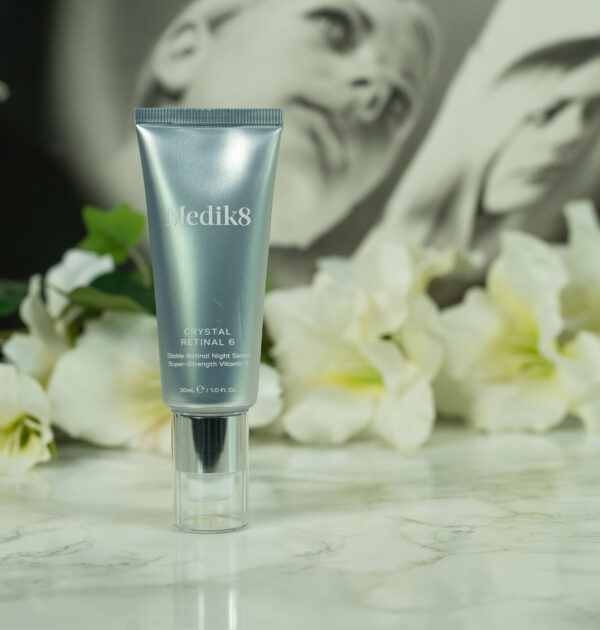 A tube of Medik8 Crystal Retinal 6, silver and with a pump, standing in front of a dark background with white flowers