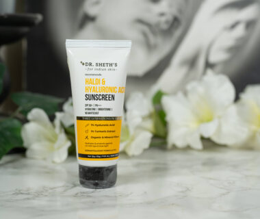 A tube of Dr Sheth's Haldi & Hyaluronic Acid Sunscreen SPF 50 PA+++ standing in front of a dark background with white flowers