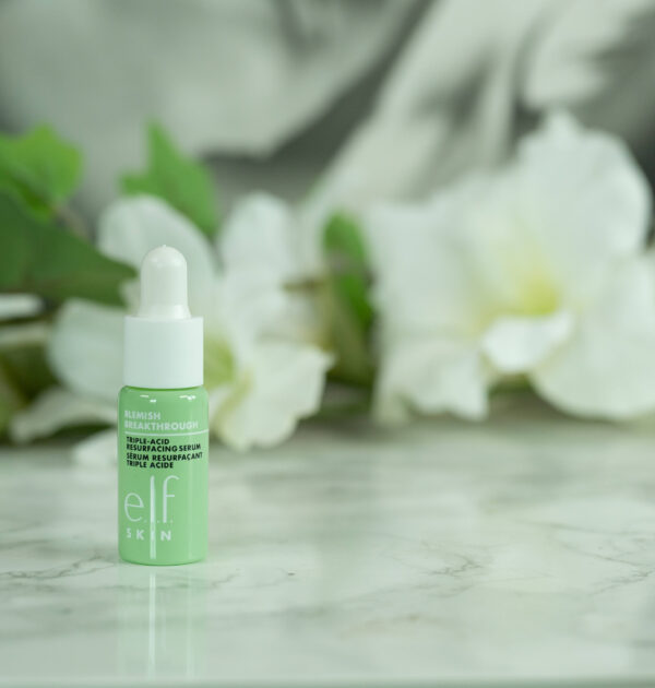 A travel size of the e.l.f. Blemish Breakthrough Triple Acid Resurfacing Serum standing in front of a dark background with white flowers