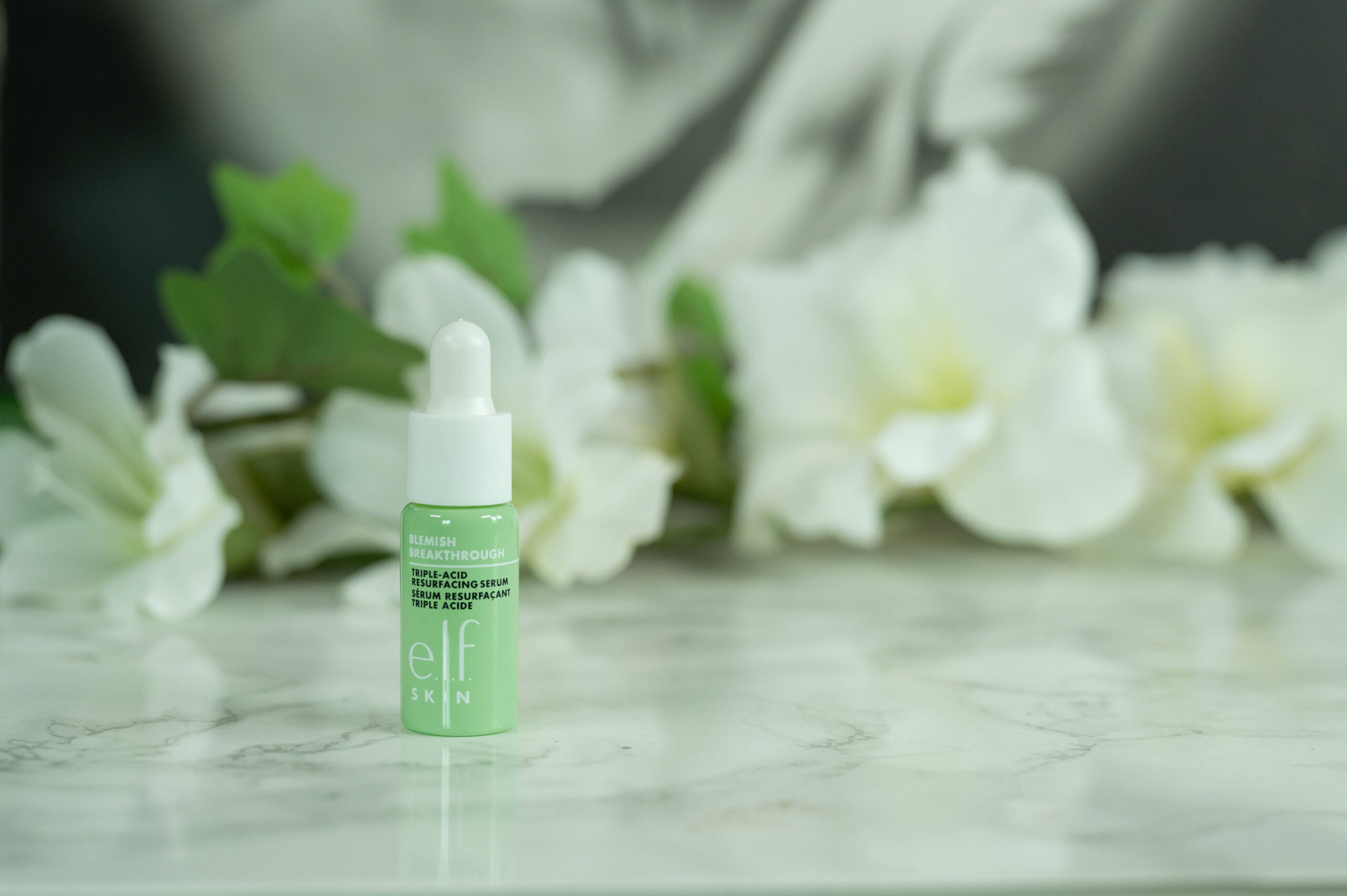 A travel size of the e.l.f. Blemish Breakthrough Triple Acid Resurfacing Serum standing in front of a dark background with white flowers