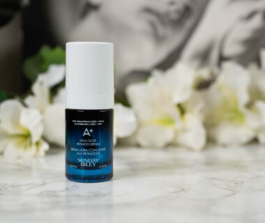 A bottle of Sunday Riley A+ High Dose Retinoid Serum standing in front of a dark background with white flowers
