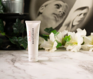 A bottle of Dr Sam's Flawless Moisturiser Intense standing in front of a dark backround with white flowers