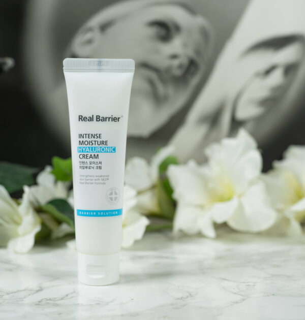 A tube of Real Barrier Intense Moisture Hyaluronic Cream (white and light blue) standing in front of a dark background with white flowers