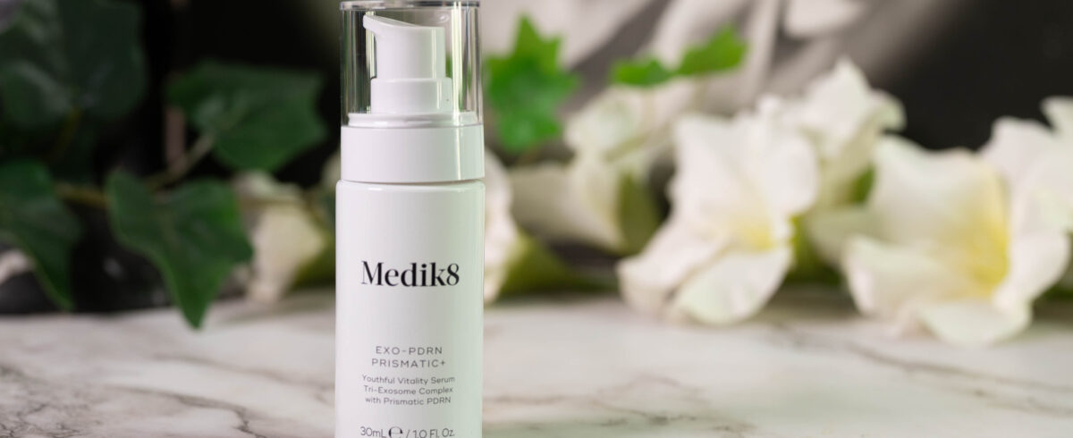 A bottle of Medik8 Exo-PDRN Prismatic+ Youthful Vitality Serum, sleek white packaging with a pump, standing in front of a dark background with white flowers.