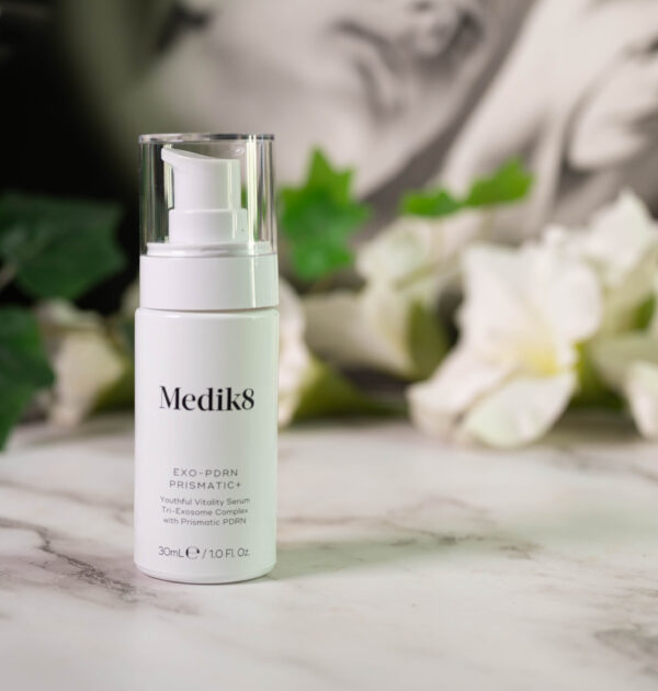 A bottle of Medik8 Exo-PDRN Prismatic+ Youthful Vitality Serum, sleek white packaging with a pump, standing in front of a dark background with white flowers.